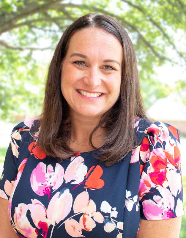 In this headhshot, Brooke Jevicks is smiling outdoors, wearing a navy blue and pink floral blouse.